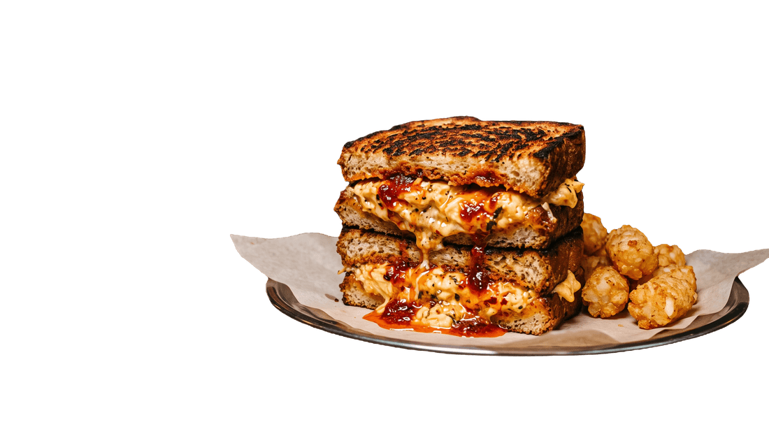 A stacked grilled sandwich oozing cheesy scrambled egg and chilli jam, with a side of tater tots, on a paper-lined tin plate